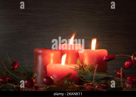 Three advent candles lit on an advent wreath with red candles in a dark room with evergreen boughs and red berries with copy space Stock Photo