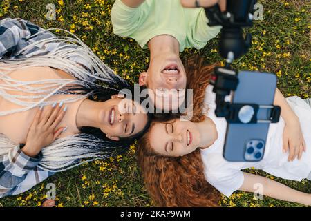 Top view portrait of smiling cheerful friends lying together on lawn taking selfie on smartphone. Top view. Multiethnic people lying on grass outdoor Stock Photo