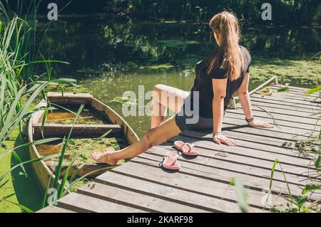 Woman sitting in sunlight on wooden bridge by old boat Stock Photo