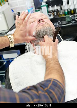 Man being shaved by a barber with an old fashioned straight razor while leaning back in a barber's chair at a barber shop. Stock Photo