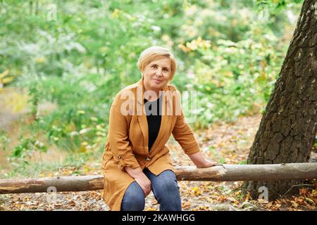 Falling season or retirement: closeup portrait of attractive caucasian senior woman sitting on a wooden log in autumn park. Smiling mature elderly people enjoying nature. High quality photo Stock Photo