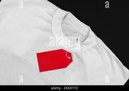White t-shirt on a black background with a red blank label. Empty red price tag on a white t-shirt. Stock Photo