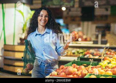 Portrait of happy and smiling hispanic female shopper in supermarket, woman smiling and looking at camera chooses apples and puts in eco bag. Stock Photo