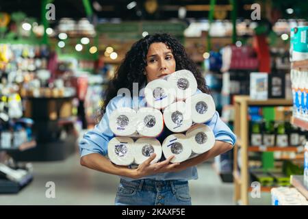 Frightened woman shopper in supermarket buys toilet paper, Latin American woman in crisis and lack of goods chooses scarce goods. Stock Photo