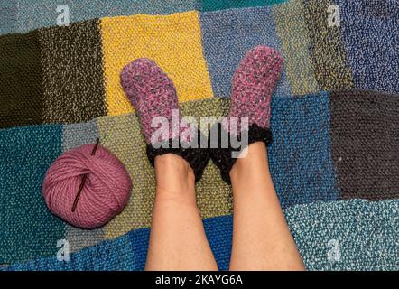 Woman feet with handmade woolen slippers or socks next to the ball of wool and crochet needle on a warm knitted background Stock Photo
