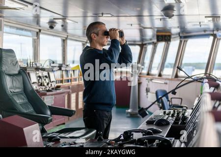 Deck officer with binoculars on navigational bridge. Seaman on board of vessel. Commercial shipping. Cargo ship. Stock Photo