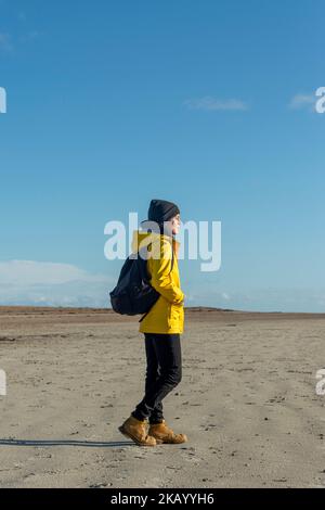 Woman wearing a yellow coat and backpack walking along a beach, Sunny autumn day. Stock Photo