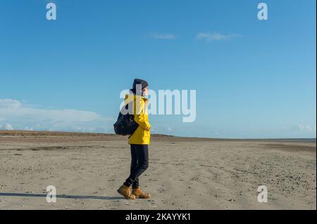 Woman wearing a yellow coat and backpack walking along a beach, Sunny autumn day. Stock Photo