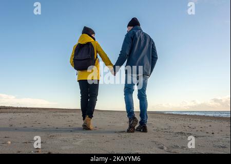 Rear view of a mature couple walking at beach holding hands, backpack and yellow coat, fall. Stock Photo