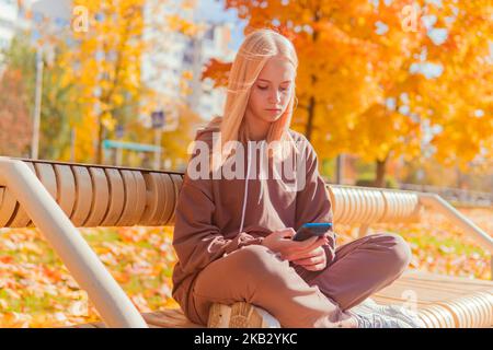 a blonde schoolgirl sits on a bench and looks at the phone against the background of yellow autumn trees. Stock Photo