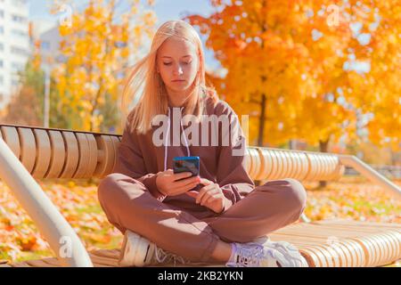 a blonde schoolgirl sits on a bench and looks at the phone against the background of yellow autumn trees. Stock Photo