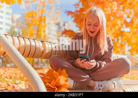 a blonde schoolgirl sits on a bench and looks at the phone against the background of yellow autumn trees. Stock Photo