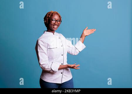 Smiling professional cook in kitchen uniform holding imaginary product in front of camera. Female kitchen staff member showing an advertising space with arms raised, studio shot with blue background. Stock Photo