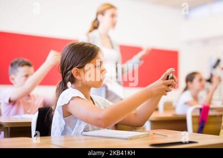 Schoolgirl using phone in classroom during lesson Stock Photo