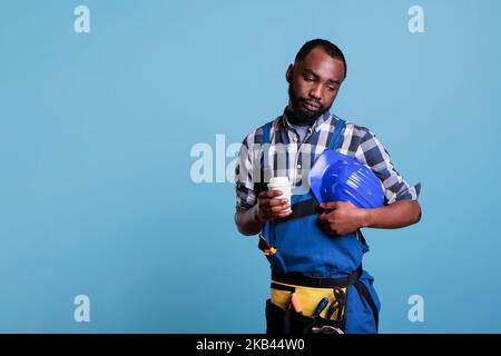 Bored construction employee drinking coffee during break against blue background in studio shot. African american handyman holding protective helmet wearing work uniform while resting. Stock Photo
