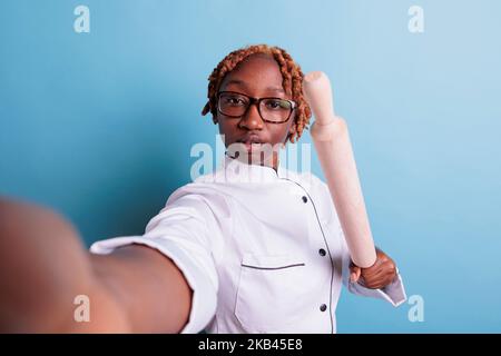 Angry female dinning hall subordinate threatening via video call. Annoyed african american chef intimidating with rolling pin. Self portrait of angry cooker in uniform. Stock Photo