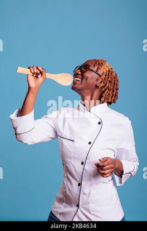 African american woman chef happily posing with ladle vertical shot. Stove member singing happily, contentedly holding kitchen utensil. Afro female cooker using glasses portrait. Stock Photo