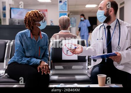 Healthcare professional receiving african american patient in hospital room. Doctor attending sick woman in clinic waiting area. Medical staff, people wearing masks at sanatorium reception. Stock Photo