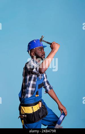 African american construction worker on ladder using hammer to drive nails into wall. Handyman wearing coveralls and protective helmet isolated on blue background in studio shot. Stock Photo