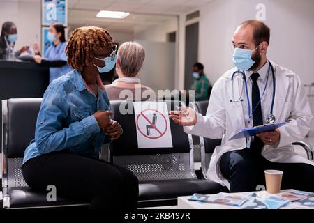 General medical practitioner explaining condition, illness to young african american female patient in hospital patient room. Physician attending sick woman, wearing cover masks for disease prevention. Stock Photo