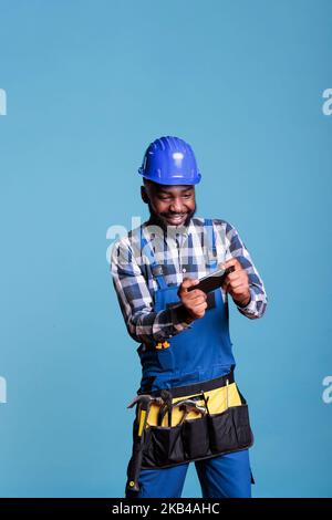 Contractor playing video games with mobile phone against light blue background in studio shot. Construction worker wearing coveralls and tool belt relaxing during break time from work. Stock Photo