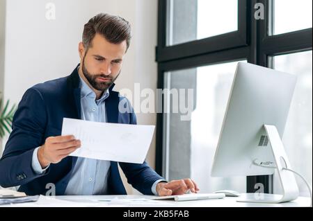 Office work. Successful confident caucasian businessman, in a suit, sits at a desk, works in a computer and with documents, looks through financial reports, analyzes the company's financial profit Stock Photo