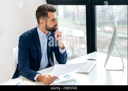 Pensive concentrated successful caucasian business man, company ceo, financial director, sits at a work desk in the office, looks at computer screen, analyzes and develops business financial strategy Stock Photo