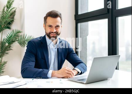 Portrait of a positive handsome confident successful caucasian man in a suit, IT specialist, investor, ceo, sits at a workplace in a modern office, looks at the camera, smiles friendly Stock Photo