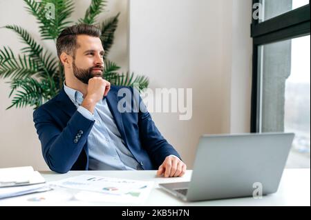 Pensive dreamy caucasian attractive successful businessman, sit at work desk, taking break from work, looking out the window, dreaming about vacation, thinking about a new project, planning a strategy Stock Photo