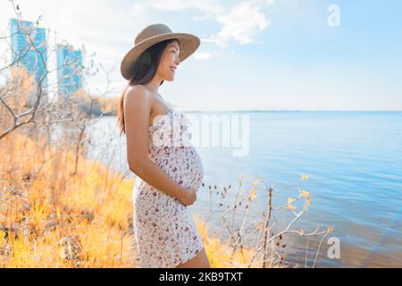 Pregnant Asian woman walking in city park by the lake wearing sun hat during spring nature walk relaxing lookig at landscape. Happy pregnancy outdoor Stock Photo