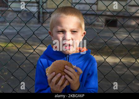 The boy raises his face from the chezburger on the background of the volleyball fence Stock Photo