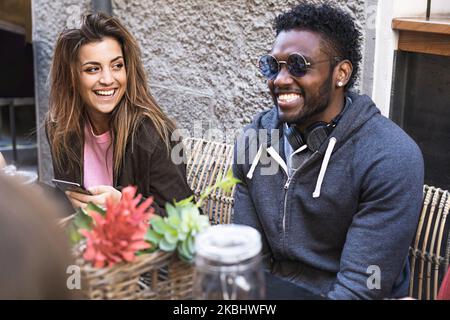 multiracial best friends laughing sitting at table - young man and a woman of different ethnicities discussing together having fun - multiethnic commu Stock Photo