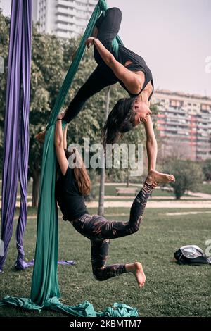 Young caucasian women practicing some aerial silks difficult figures together hanging on a bridge in a city park. Stock Photo
