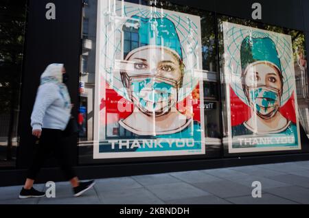 A woman wearing a face mask walks past digital displays supporting health workers in the windows of clothing retailer Flannels on a near-deserted Oxford Street in London, England, on May 6, 2020. Britain is now well into its seventh week of coronavirus lockdown, although a slight easing of the restrictions is expected to be announced this Sunday, along with a schedule for further 'easements' over the coming months. Covid-19 deaths across the country have meanwhile reached 30,076 according to today's figures from the UK's Department of Health and Social Care. The figure is currently considered  Stock Photo