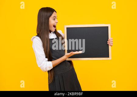 Teenager younf school girl holding school empty blackboard isolated on yellow background. Portrait of a teen female student. Excited face, cheerful Stock Photo