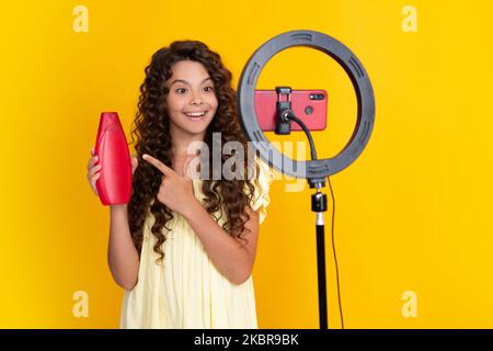 Young blogger child hold hair conditioner or shampoo bottle recording video for blog. Teen girl speaking in front of camera for vlog. Teenager working Stock Photo