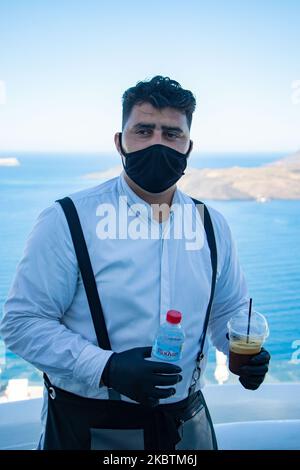 Portrait of a waiter serving the a la carte breakfast at the restaurant section with tables having distances between the tourists, instead of an open buffet. The staff is wearing a face mask, gloves and uses hand sanitizer due to Covid-19 Coronavirus pandemic safety measures in Volcano View Hotel Santorini, a 5 star hotel at Thera, Santorini Island, Cyclades, Aegean Sea in Greece. Greek government relaunched the summer tourist season, by easing the traffic and travel ban measures, the lockdown and quarantine, but also testing the arriving passengers. Tourism a very important sector for the cou Stock Photo