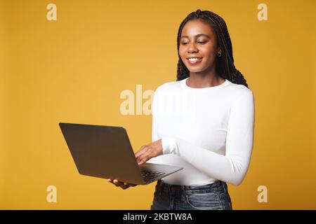Confident modern woman using laptop isolated on yellow. Multiracial female entrepreneur typing while studying online Stock Photo
