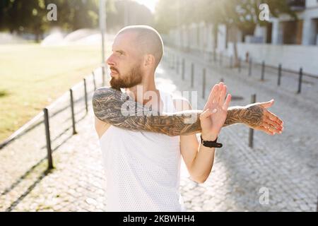 Young muscular tattooed man doing exercises, stretching early in the morning, bald bearded guy in sportswear stretching arms before jogging Stock Photo