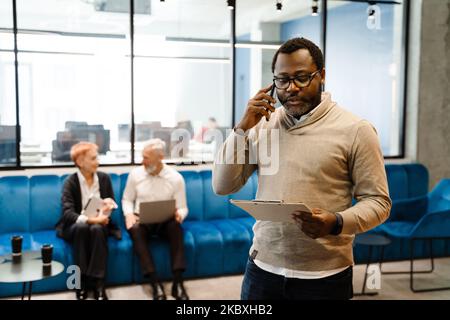Bearded black man talking on cellphone while working with colleagues in office Stock Photo
