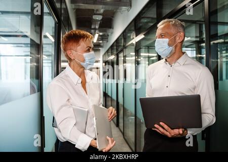 Two white colleagues in face masks talking while working in office Stock Photo
