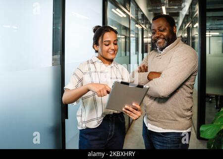 Two multiracial colleagues talking and using tablet computer while working in office Stock Photo