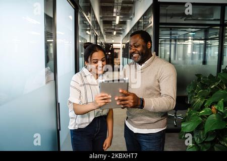 Two multiracial colleagues talking and using tablet computer while working in office Stock Photo
