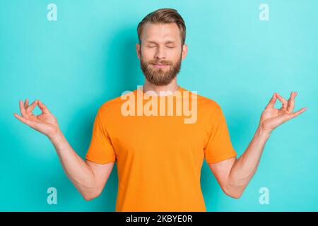 Photo of concentrated meditating good mood man with blond hairstyle dressed orange t-shirt eyes closed isolated on teal color background Stock Photo
