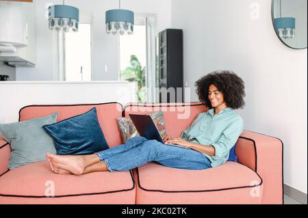 Full length view of the black female businesswoman or female freelancer in comfy casual clothes sitting on the couch and working on the project, typing email on the laptop, writing an article Stock Photo