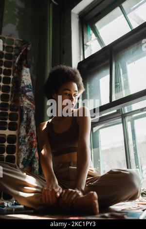 pensive african american woman sitting on windowsill with crossed legs and looking through window in art workshop Stock Photo