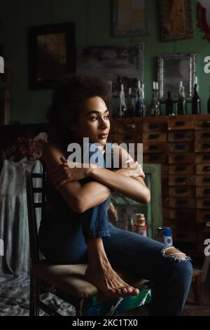 pretty and thoughtful african american woman in overalls sitting with crossed arms in art studio Stock Photo