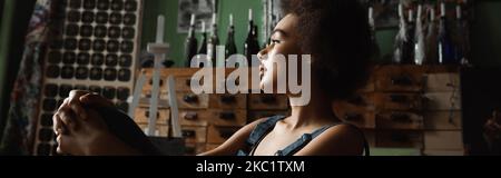 profile of thoughtful african american woman sitting in workshop and looking away, banner Stock Photo