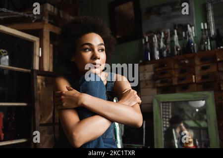 pensive african american woman sitting with crossed arms in art studio near vintage mirror on blurred background Stock Photo