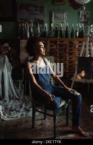 brunette african american woman in overalls sitting in workshop with vintage decor and looking away Stock Photo